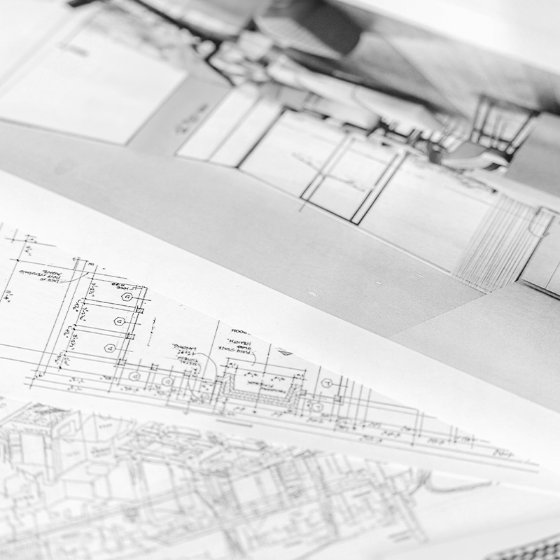 Black and white image showing close-up views of architectural blueprints and technical drawings laid out on a table, capturing detailed floor plans and structural diagrams. The image highlights the intricacy and precision involved in architectural design.