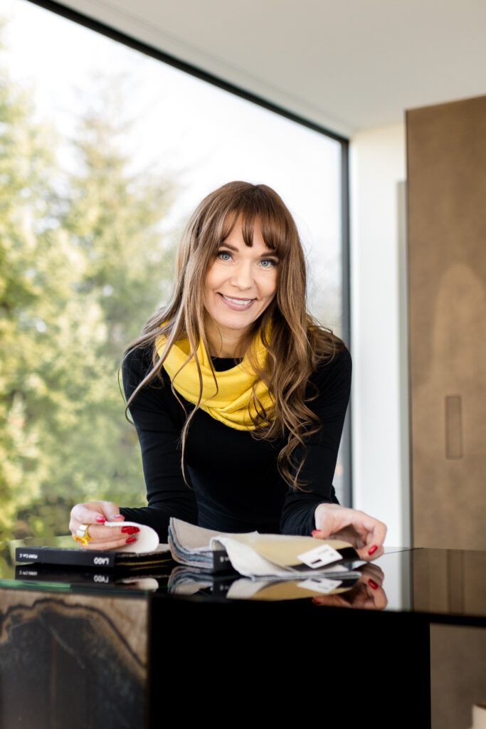 A woman with long brown hair and bangs is smiling while standing at a table with fabric swatches and design tools. She is wearing a yellow scarf and black top. A large window in the background shows a blurred view of trees outside.